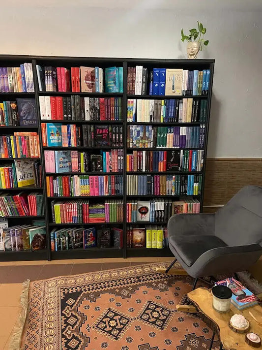 Cozy reading nook with black bookshelves filled with colorful books, a gray armchair, a patterned rug, and a small wooden table with candles and books.