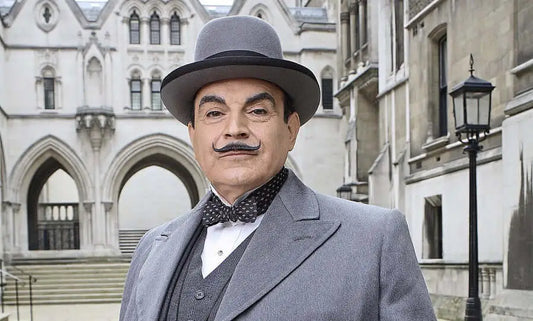 Man in grey suit and bowler hat with mustache, standing outside historic building.