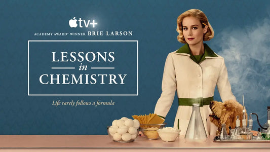 Elizabeth Zott, portrayed by Brie Larson, stands confidently in a bright kitchen wearing a 1950s-style red dress and apron, holding a mixing bowl and spoon. The scene reflects a retro, vibrant aesthetic from the TV adaptation of “Lessons in Chemistry.”