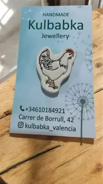 Handmade ceramic hen pin with white body, red comb, black details on blue Kulbabka card