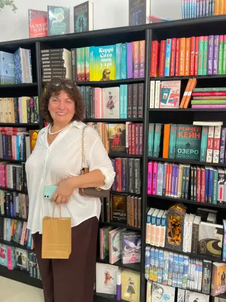 Smiling woman with brown hair in white shirt holding phone and bag, in front of bookshelves, coach Valentyna Melnyk